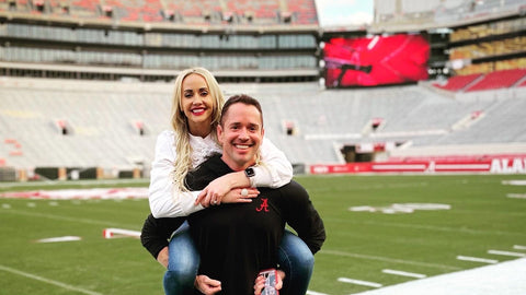 Man carrying a woman on a football field with stadium seats and advertising boards in the background.